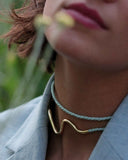 Close-up of a woman wearing a gold necklace and light blue cord around their neck with a blurred green background.