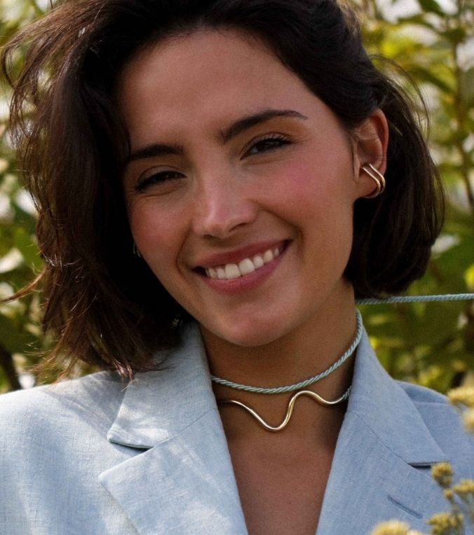 Woman wearing a light blue blazer and a metal and blue ribbon necklace with a blurred natural background