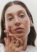 Woman wearing gold earrings and ring, touching her face against a neutral background