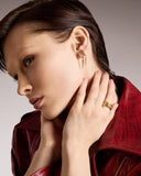 Woman wearing gold drop earrings and a ring, with a red jacket on a neutral background