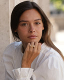 A woman with long dark brown hair wearing a white blouse and a gold geometric ring, posing with her hand near her chin against a softly blurred beige background.