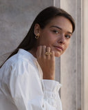 A woman wearing a white blouse and gold jewelry, including a gold hoop earring and a gold ring, posing against a textured gray wall.