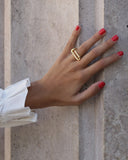 A gold geometric ring worn on a hand with red nail polish, resting against a textured stone wall.