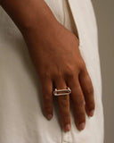 A close-up of a person's hand wearing a minimalist silver safety-pin style ring, resting against a soft off-white textured fabric background.